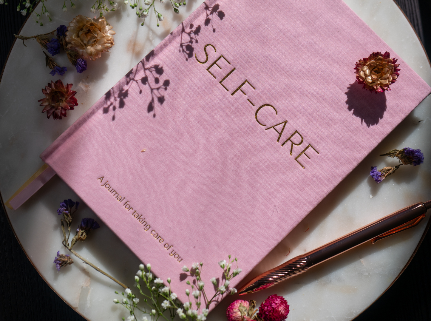 Pink 'Self-Care' journal on a marble surface with flowers